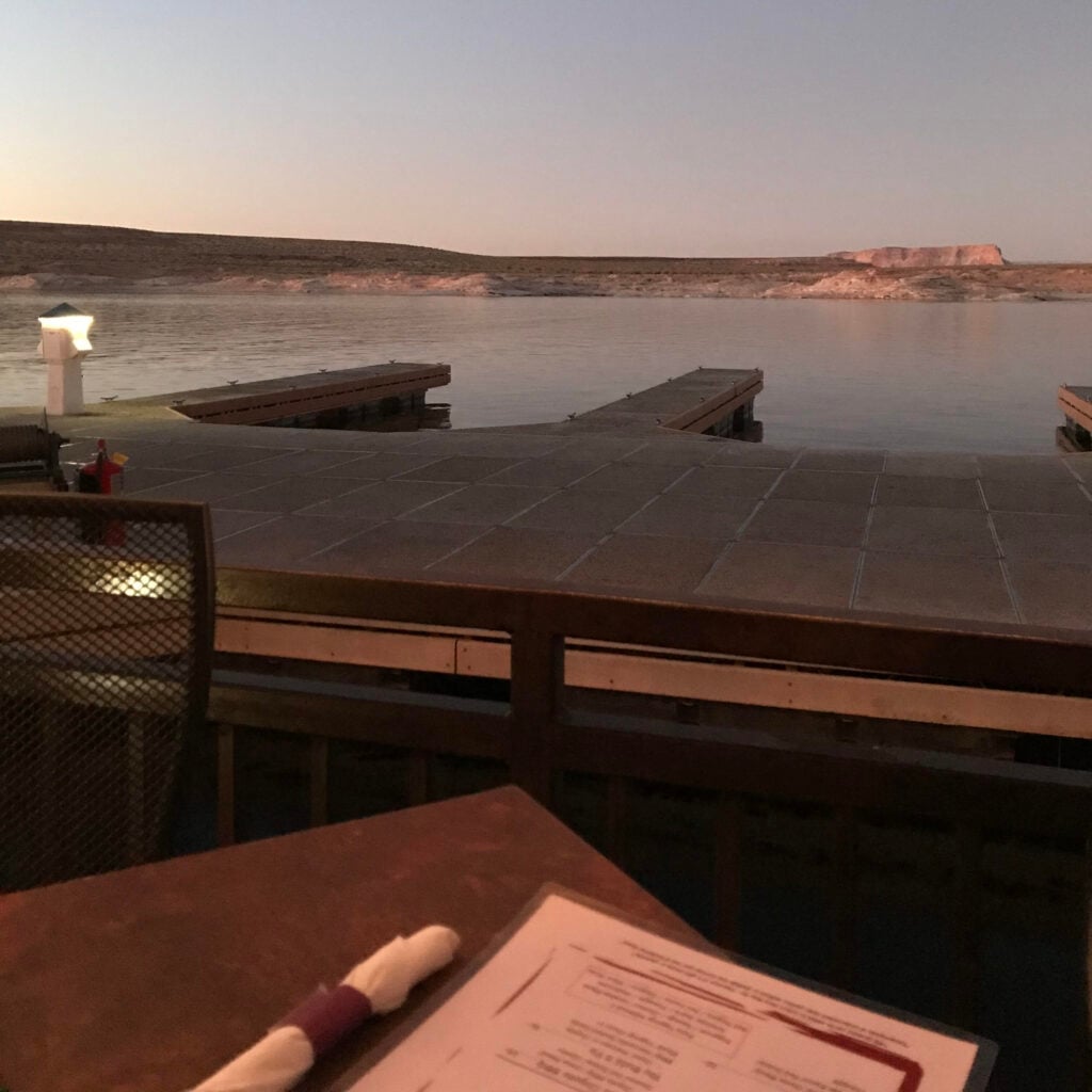 A restaurant menu sits on a table overlooking a dock and lake with sandstone hills in the background.
