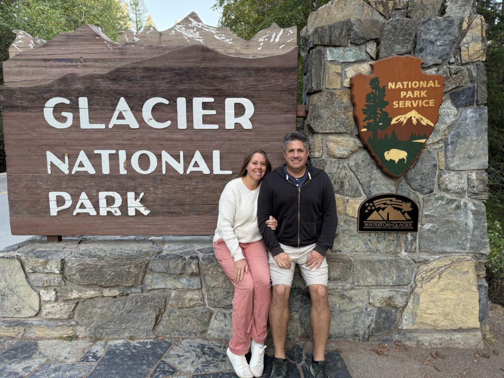 man and woman sitting at Glacier National Park entrance sign