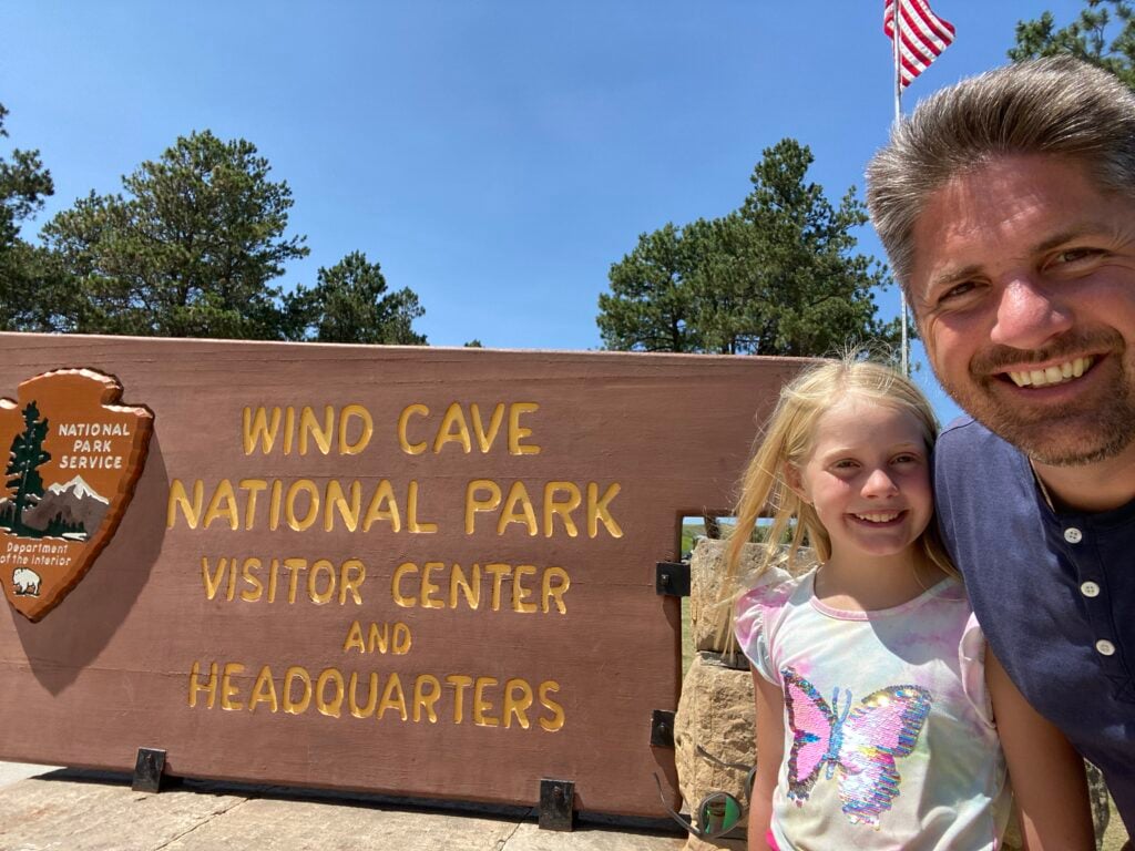 Man and child standing next to a national park sign for Wind Cave