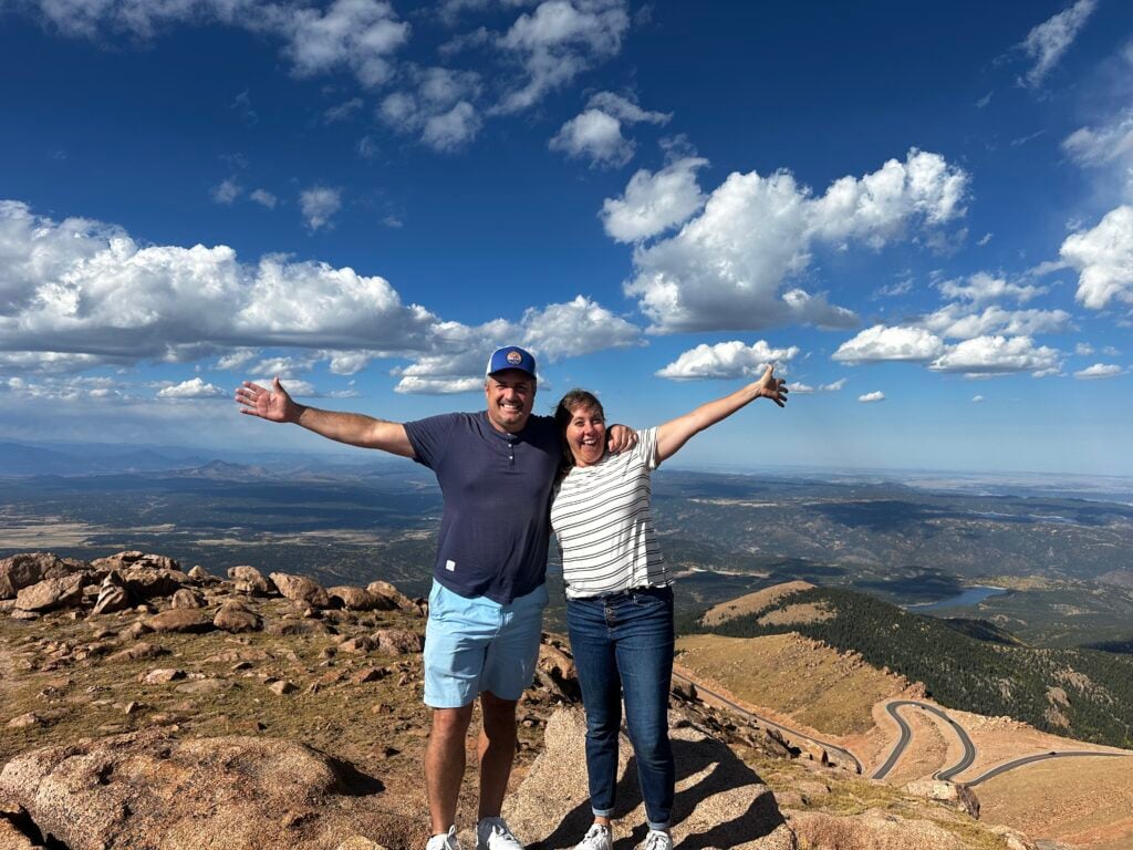 Man and woman smiling with arms in the air with a valley and winding road behind them