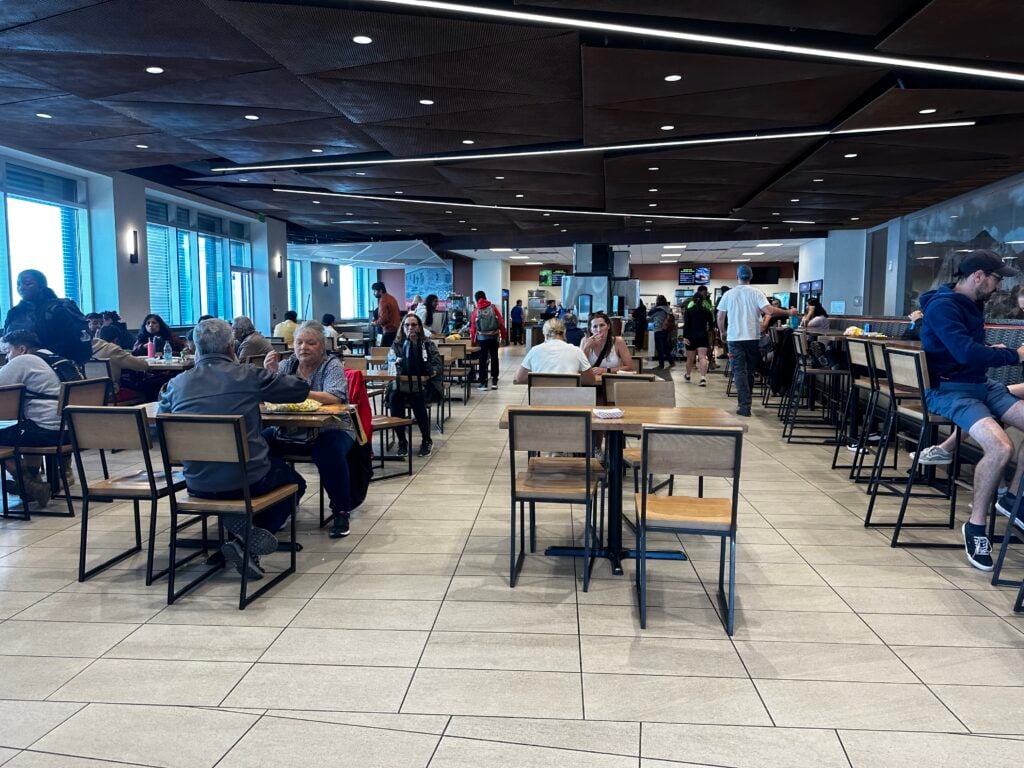 Large cafeteria with several metal and wooden chairs with close to 70 people dining