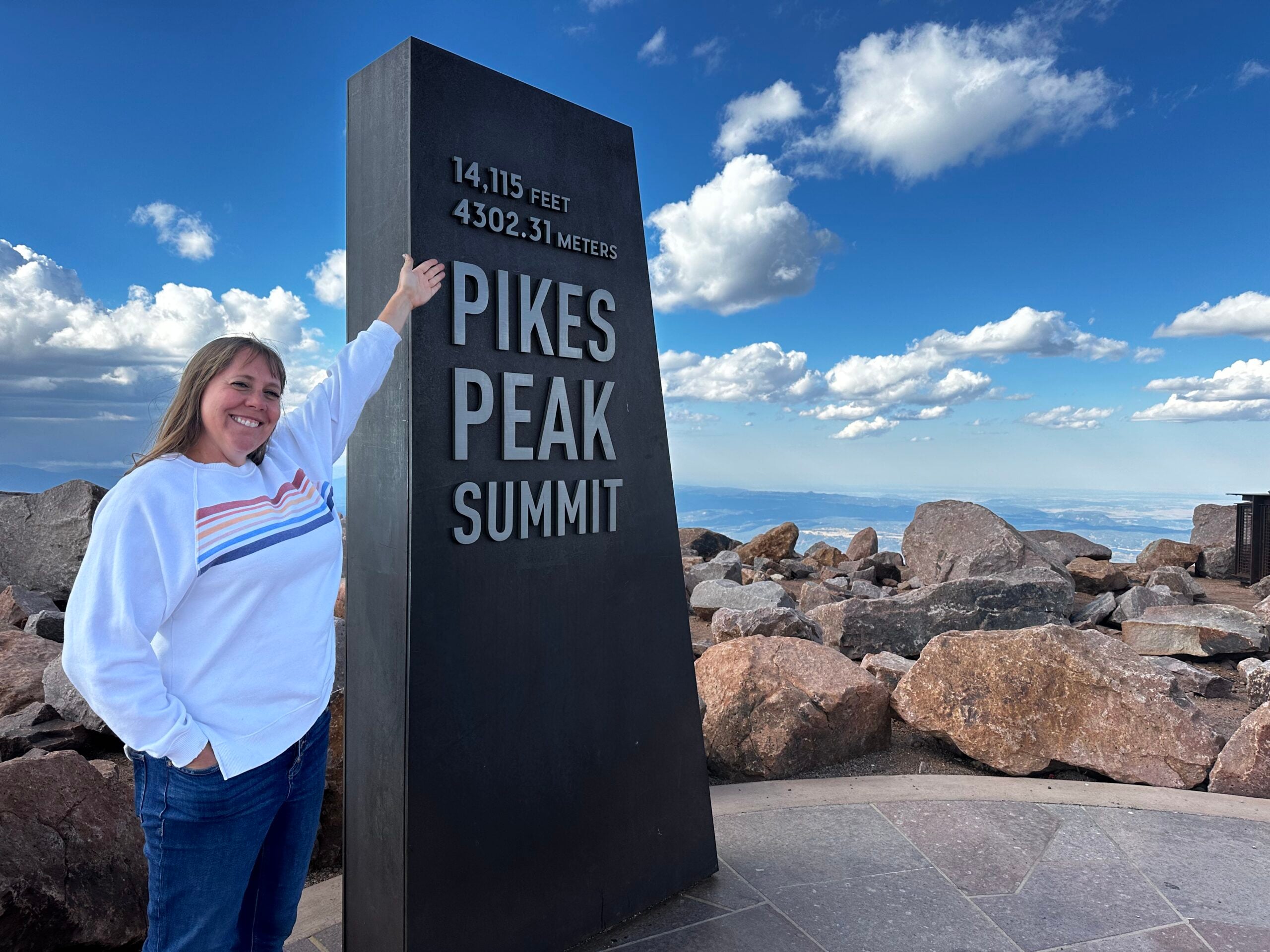 Cheryl standing beside the Pikes Peak summit sign with mountain views behind her