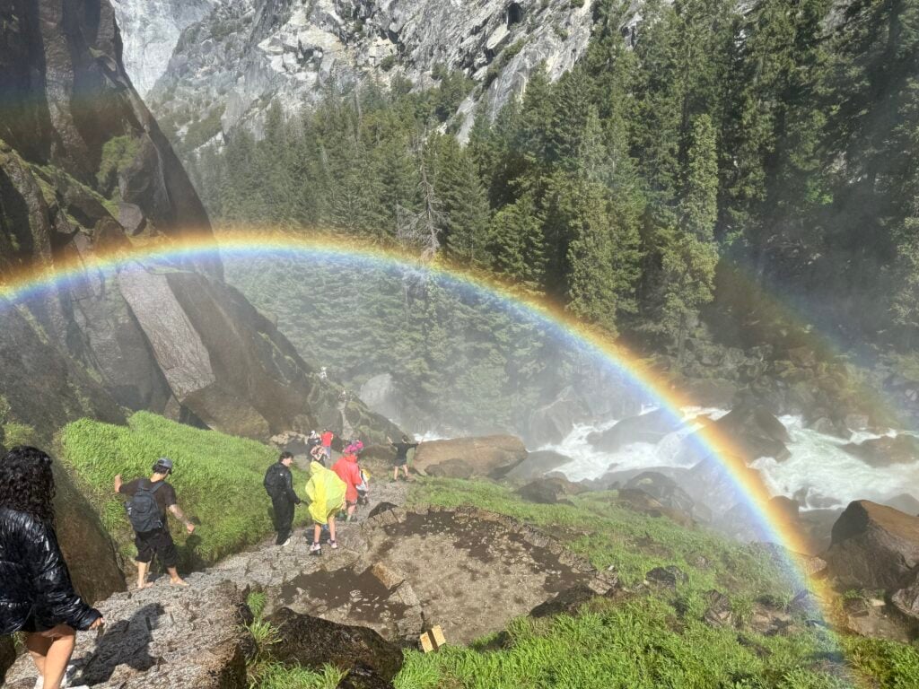 Rainbow going over people hiking on a trail next to a cascading river