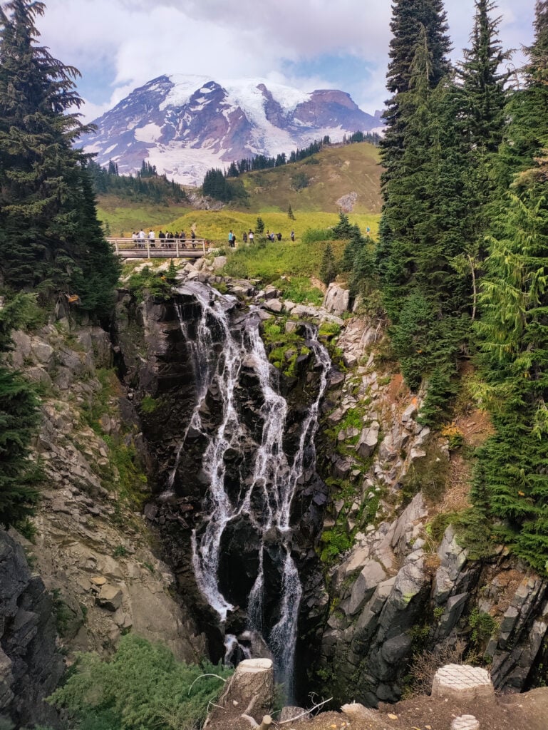 people crossing bridge over waterfall with Mount Rainier in the distance