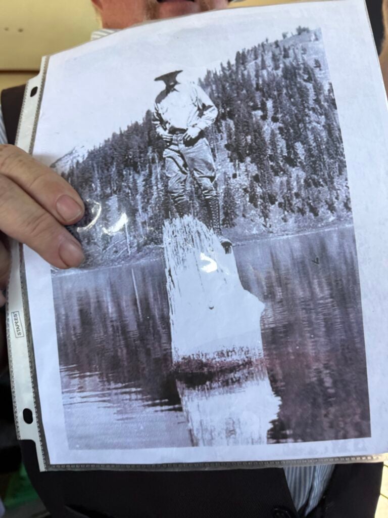 Black and white photo of man standing on a vertical log floating in Crater Lake