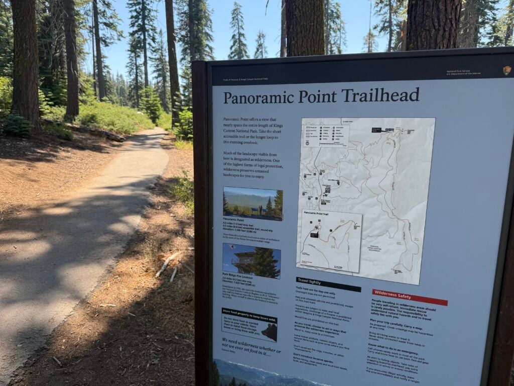 Sign with a map and paved trail going through large pine trees