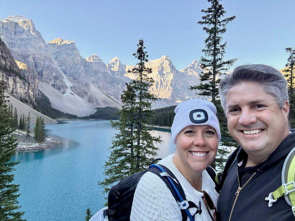 man and woman in front of glacial lake in front of glaciers and mountain peaks
