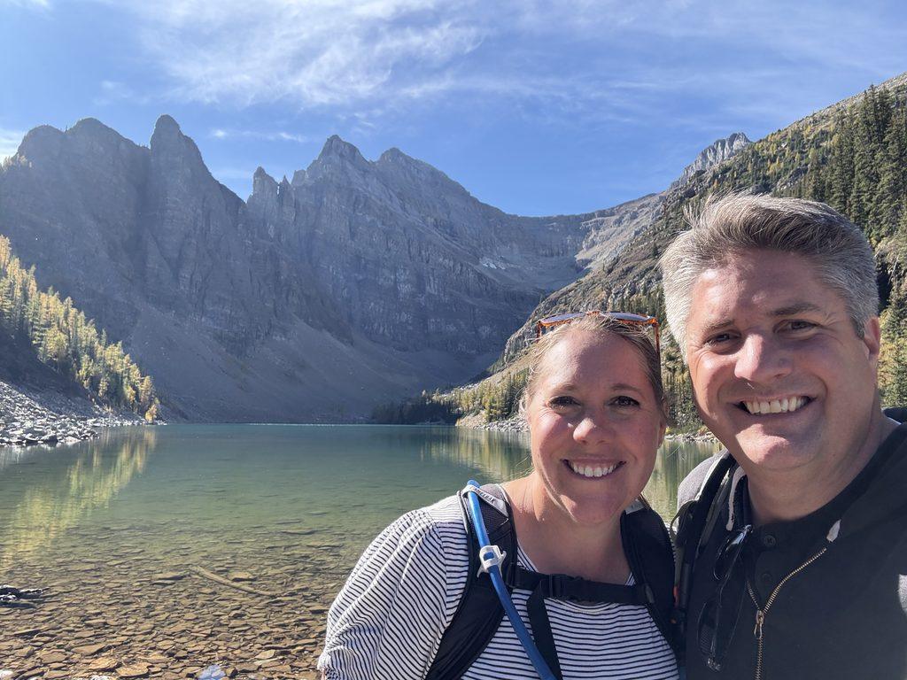 man and woman standing in front of green lake with pointed mountain peaks in the background