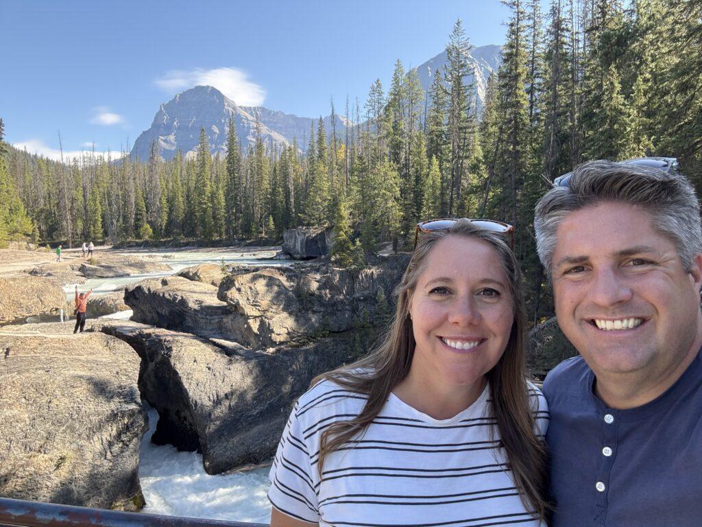 man and woman in front of river with natural bridge crossing