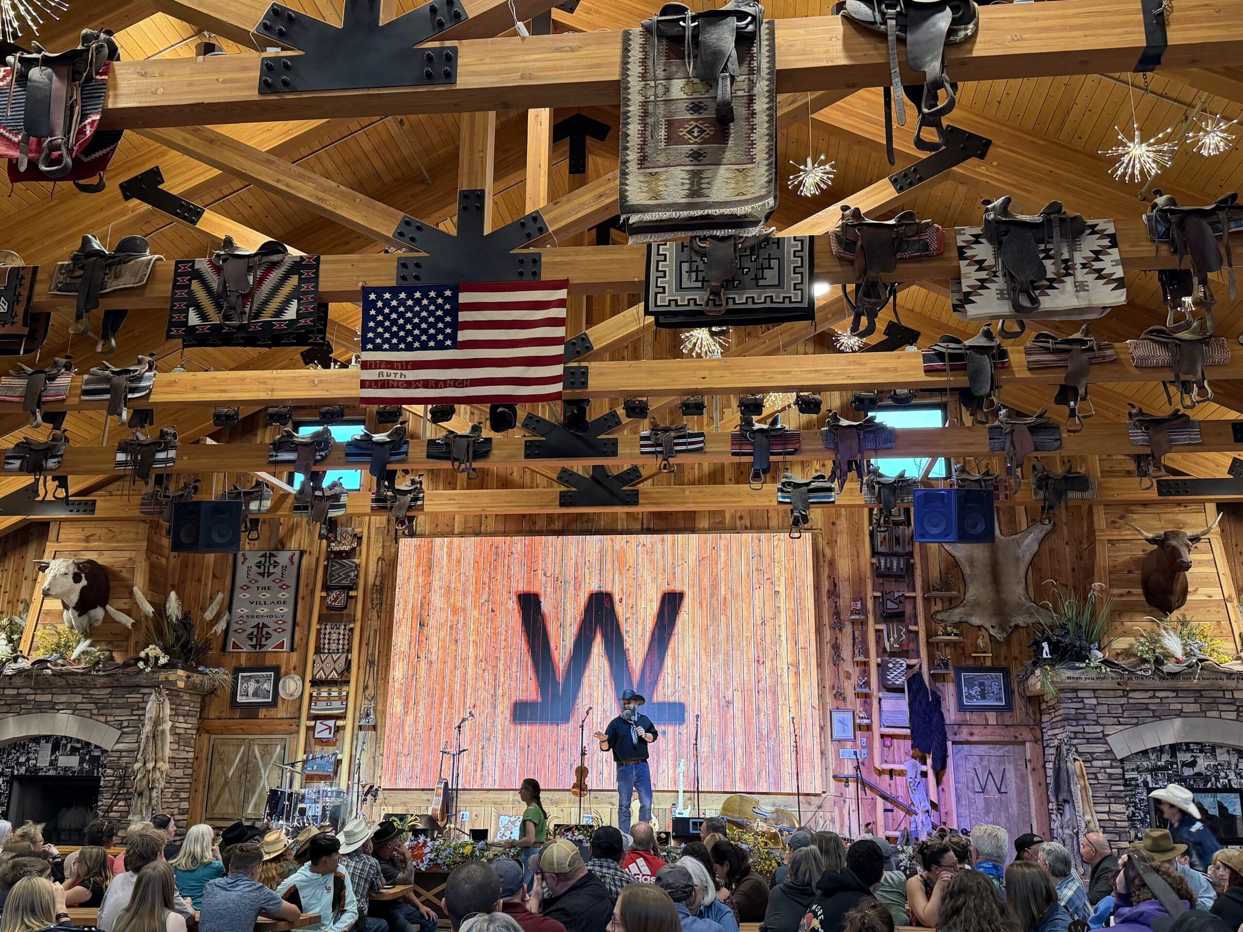 A performer on the Flying W Ranch stage beneath wooden beams, saddles, and an American flag