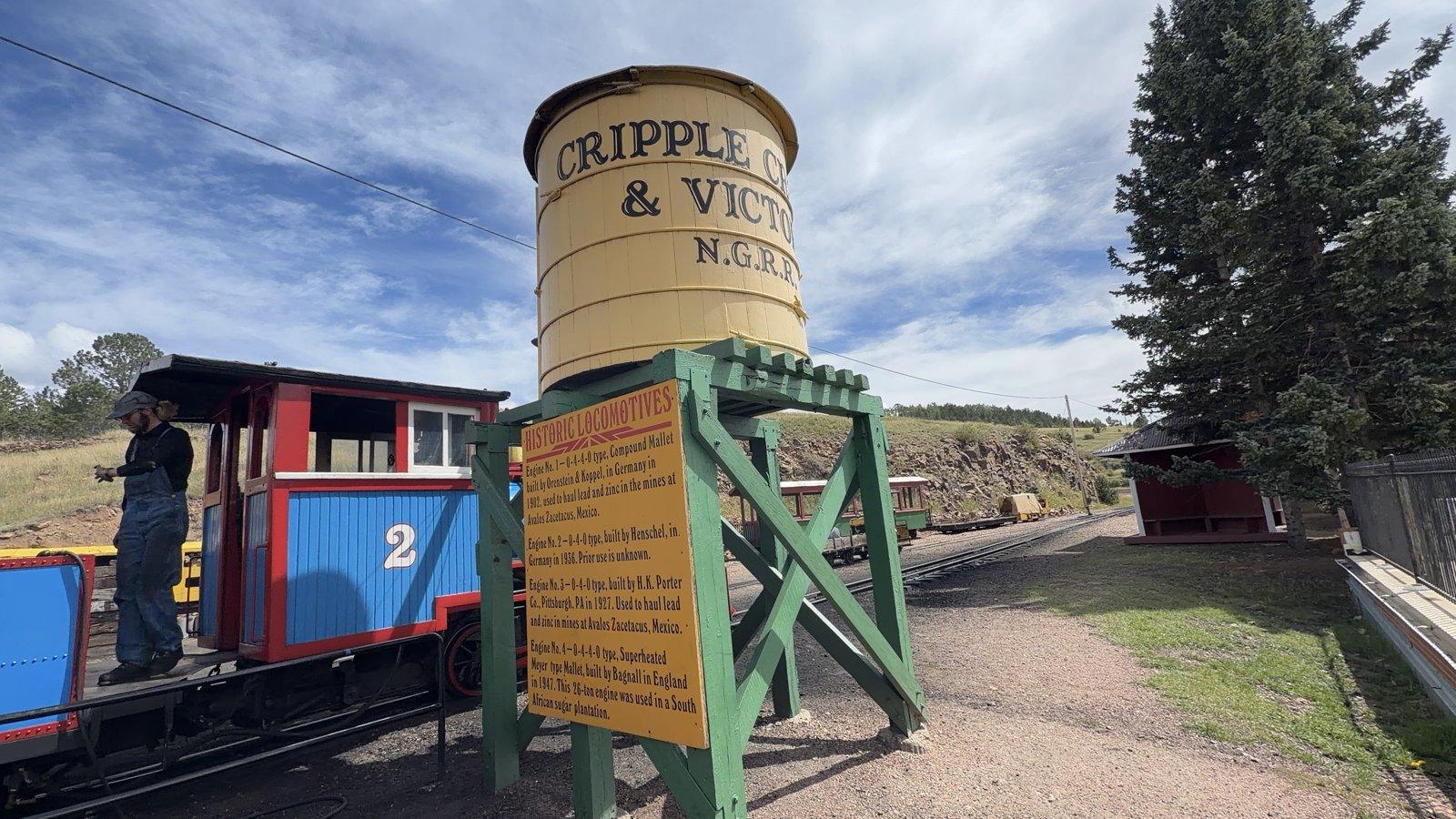 Wooden Cripple Creek and Victor railroad water tower beside the blue steam engine at the depot.