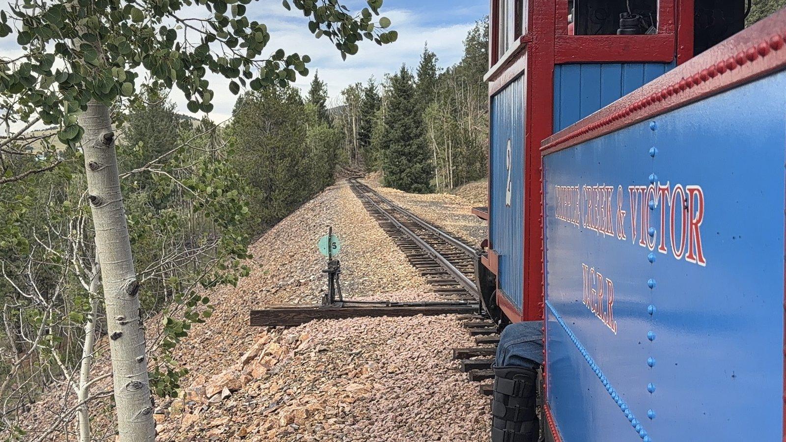 View beside the blue locomotive along narrow-gauge tracks bordered by green aspen trees.