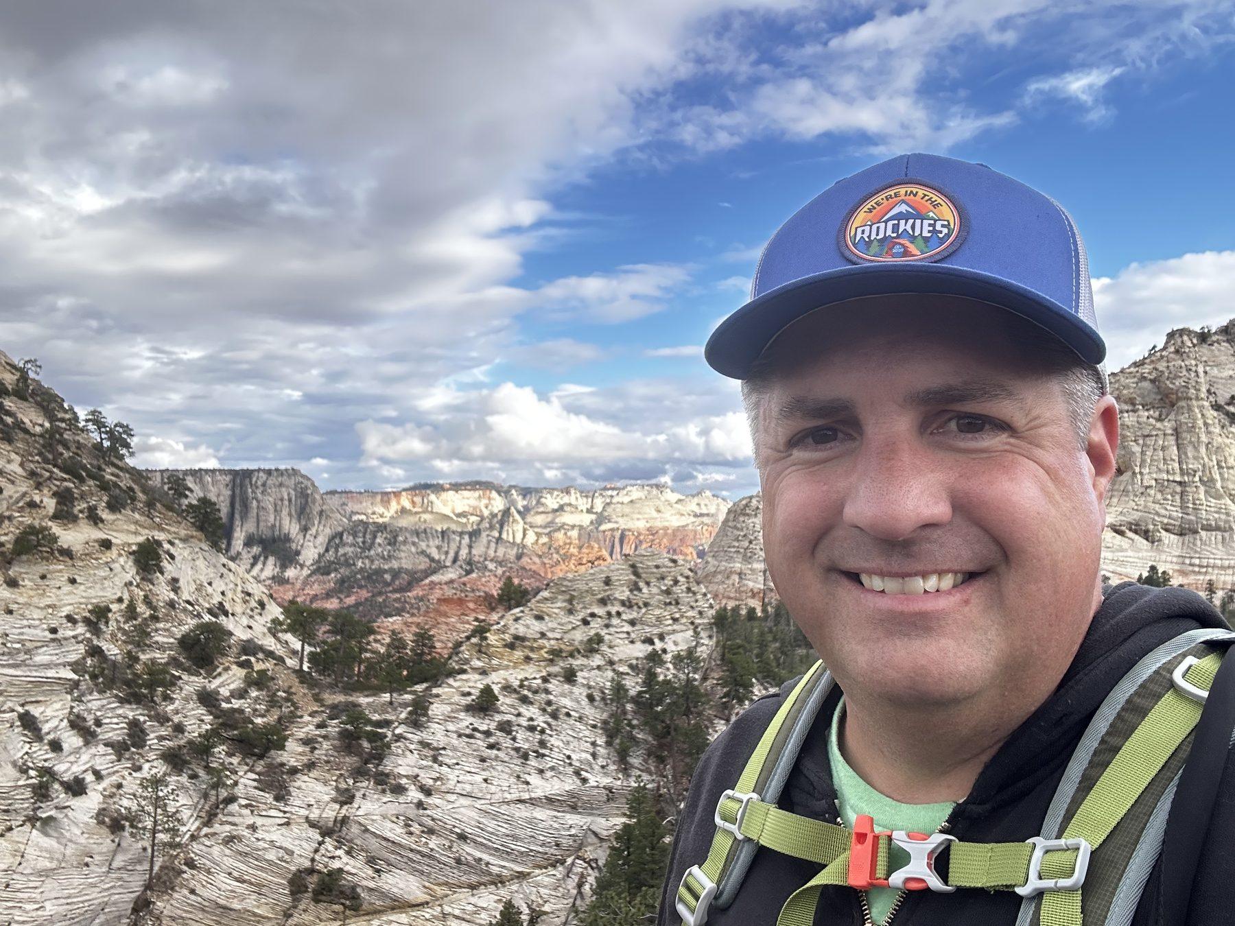 Matt smiles at the Northgate Peaks overlook with sandstone cliffs and a cloudy sky behind him.